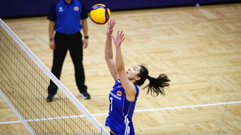 A female volleyball player jumps at the net to block a moving ball during a match.